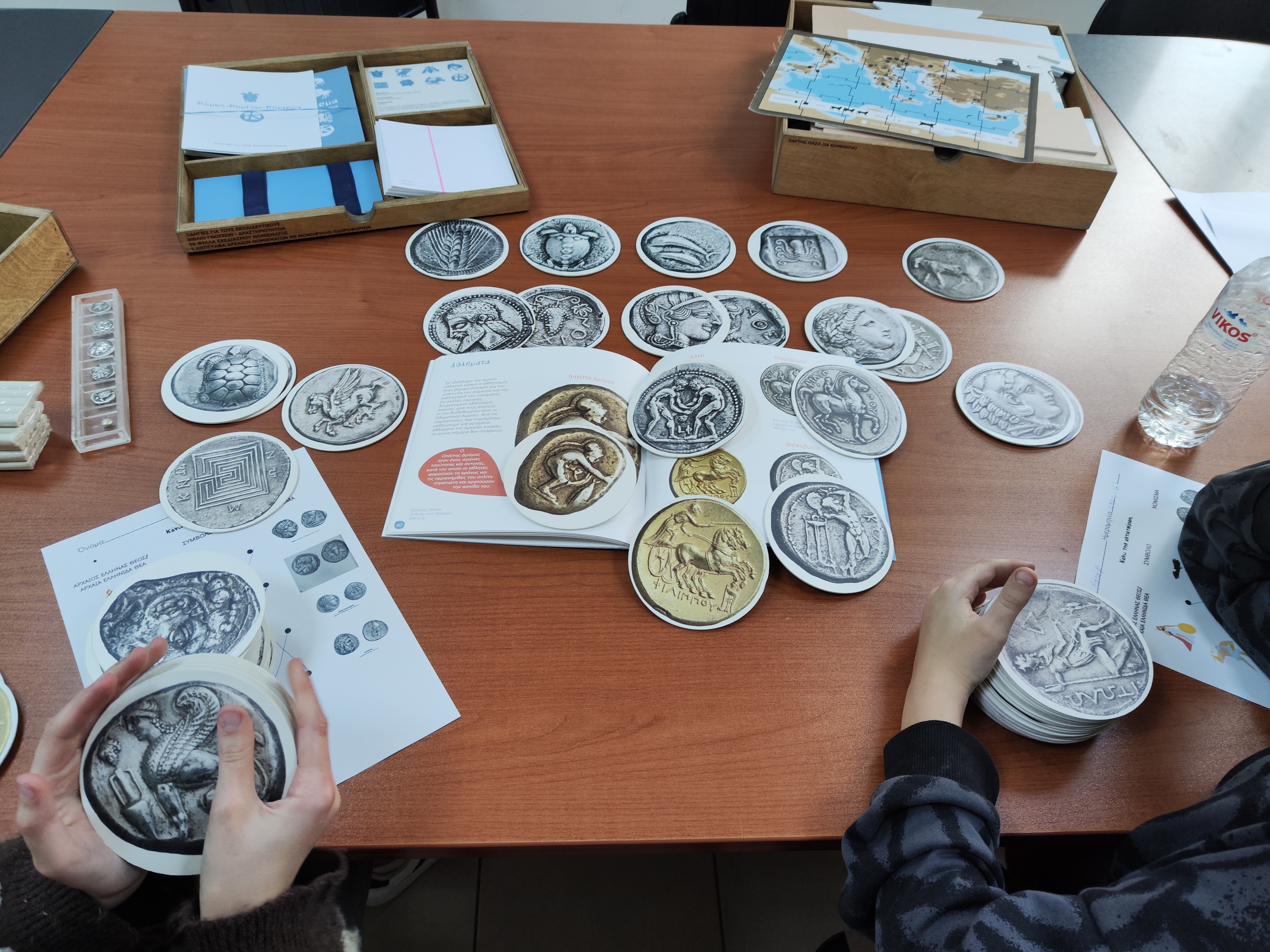 Πάνω σε τραπέζι 2 ξύλινες θήκες με εκπαιδευτικό υλικό και απλωμένα χάρτινα αντίγραφα νομισμάτων. Στιγμιότυπο από εκπαιδευτικό πρόγραμμα