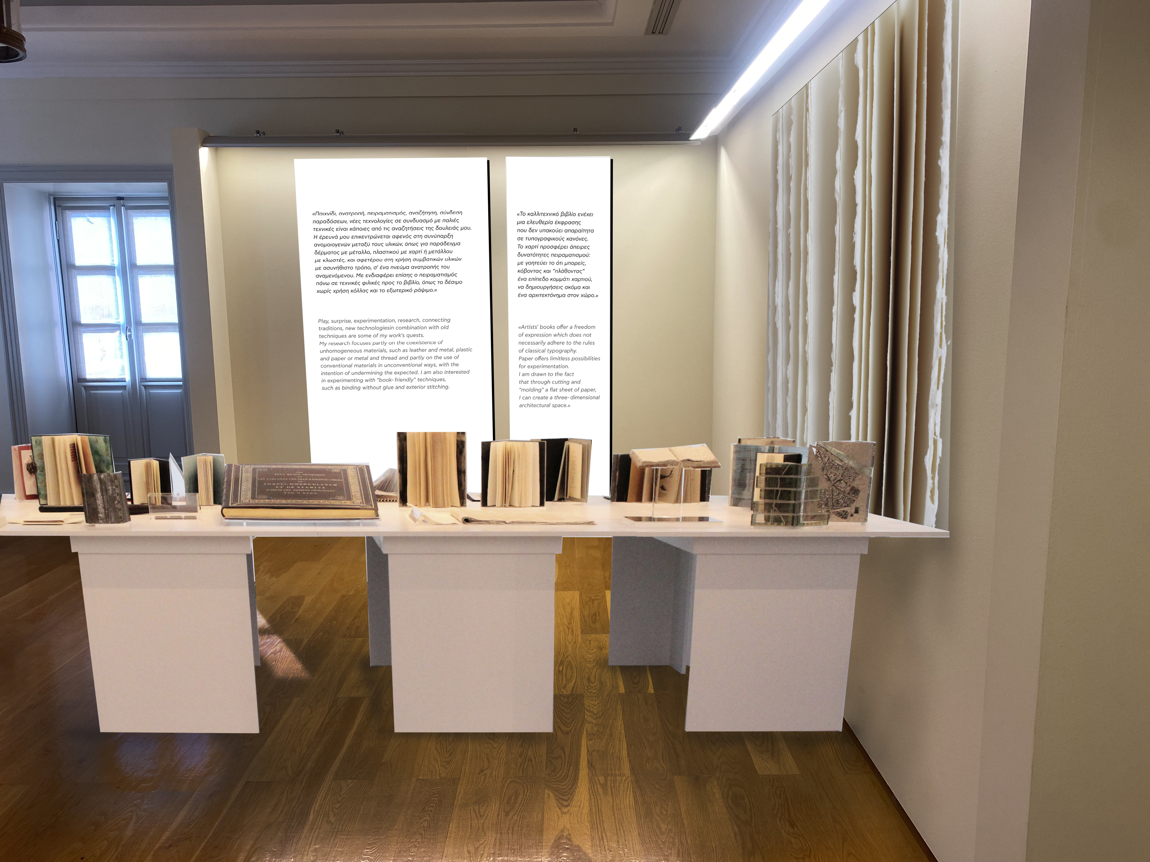 Large table with standing books. In the background, white boards inscribed with quotes by the artist.  A half-open standing book with double black cover and back cover, and white pages.