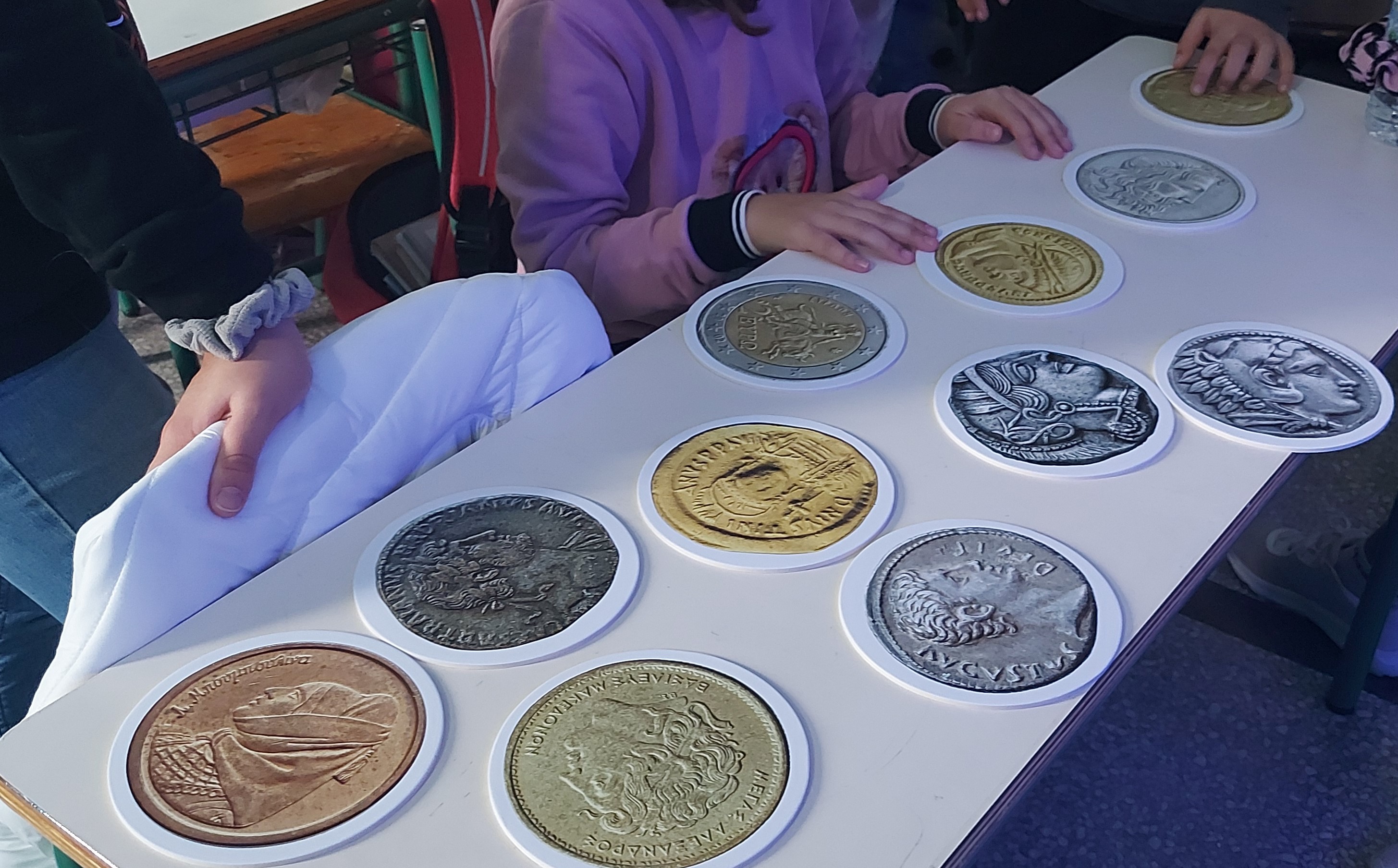 Μαθητές και μαθήτριες επεξεργάζονται χάρτινες απομιμήσεις 11 νομισμάτων που βρίσκονται πάνω σε λευκό θρανίο. Μεγεθυμένες εκτυπώσεις νομισμάτων