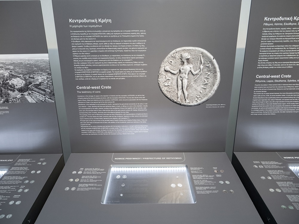 Νόμισμα της κεντροδυτικής Κρήτης σε φωτεινή προθήκη και σχετικό εποπτικό υλικό.   Έκθεμα «Κρητών πόλεις. Η μαρτυρία των νομισμάτων»
