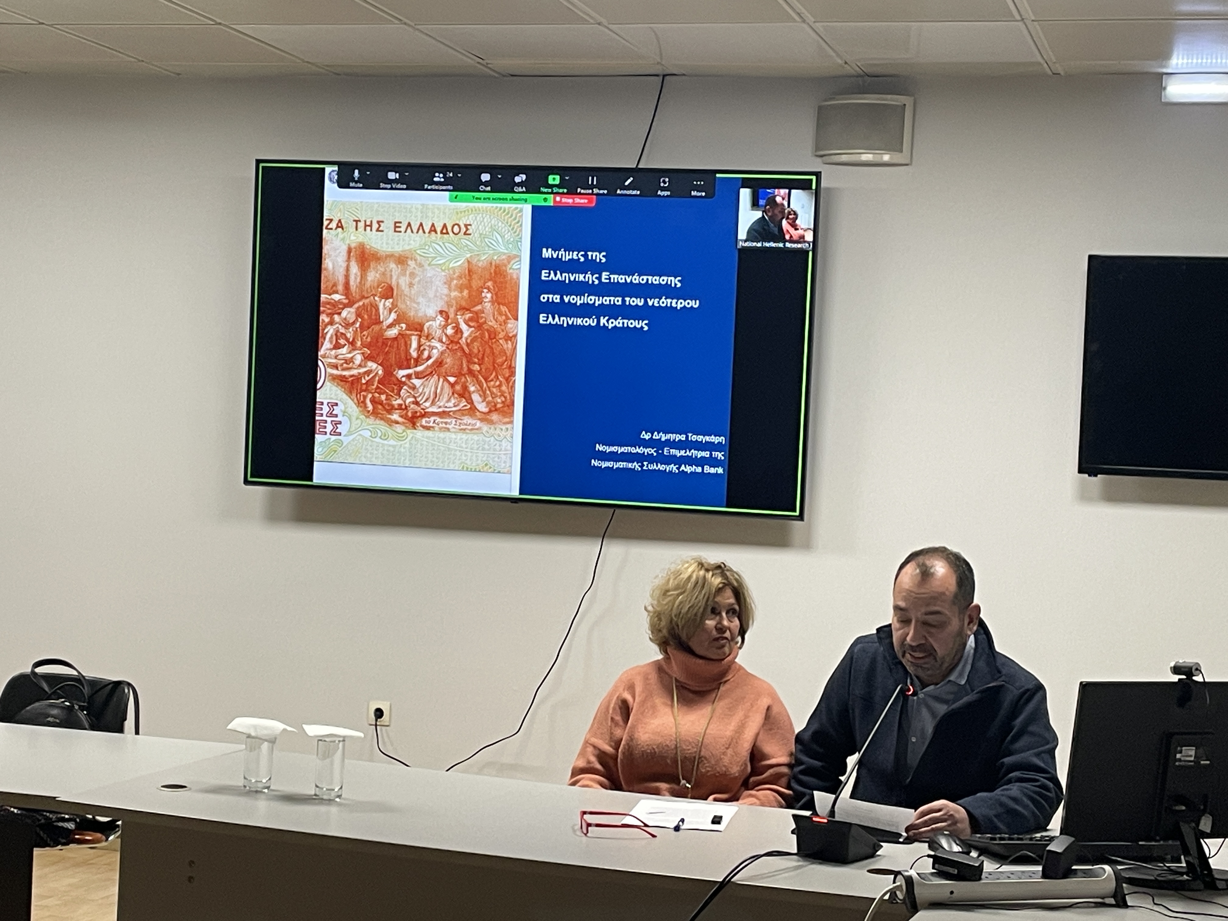 Η ομιλήτρια και ο κ. Τσέλεκας, πρόεδρος Σωματείου Φίλοι Νομισματικού Μουσείου. Πίσω οθόνη με χαρτονόμισμα. Στιγμιότυπο της διάλεξης