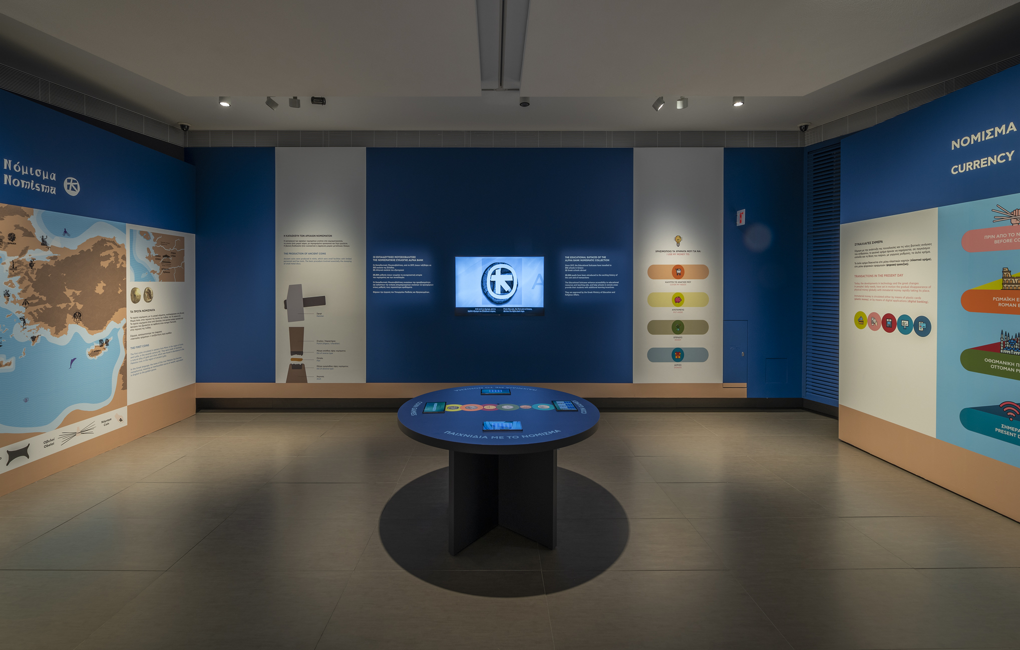 Αίθουσα με εικόνες και πληροφορίες στους τοίχους, και στο κέντρο τραπέζι με τάμπλετ.  Έκθεση «Η άλλη όψη του νομίσματος»