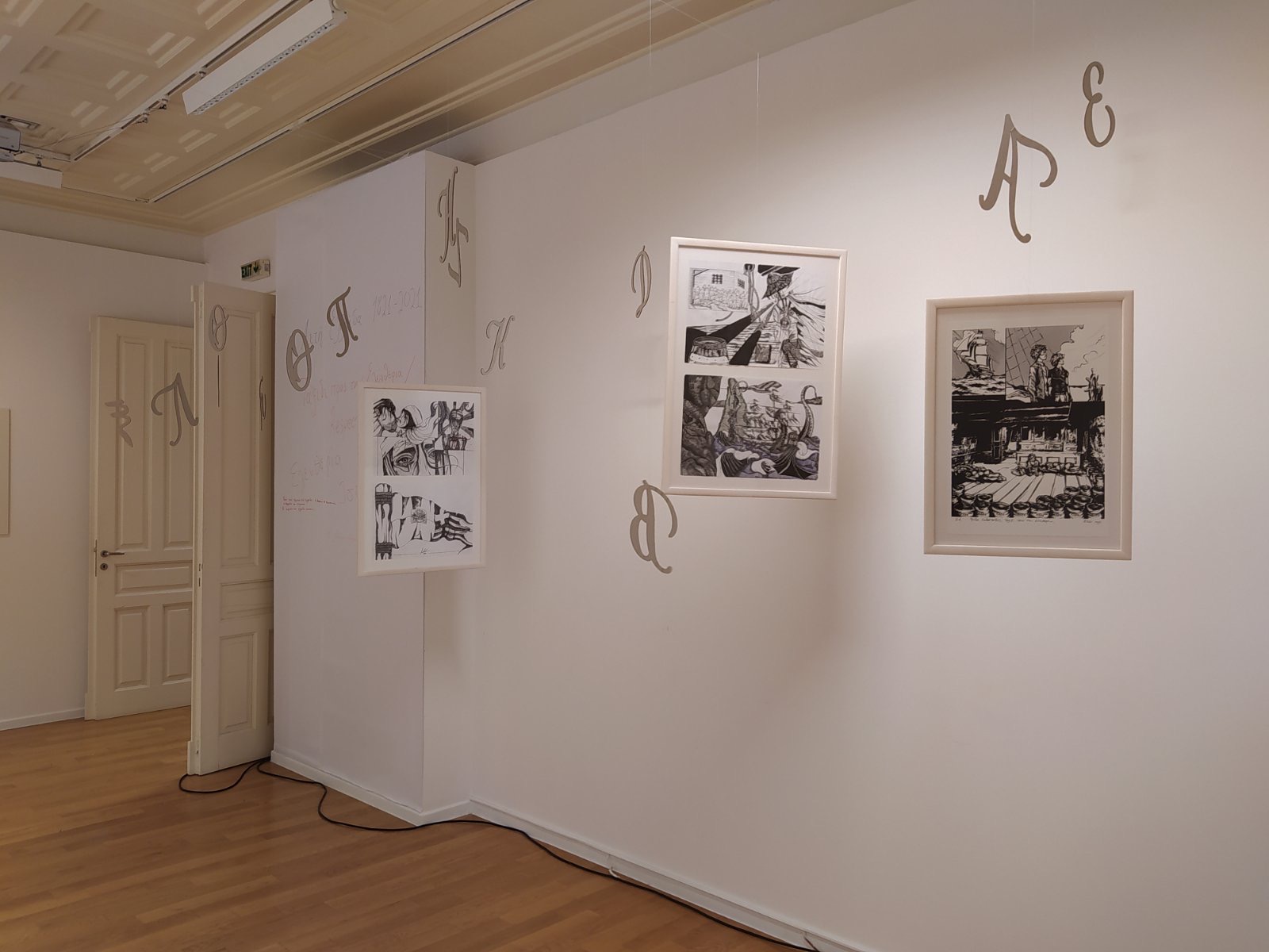 Photo of the exhibition with paintings on the walls and letters hanging from the ceiling.  “Haiti Greece 1821-2021. Voyage to Freedom/RESPECT”