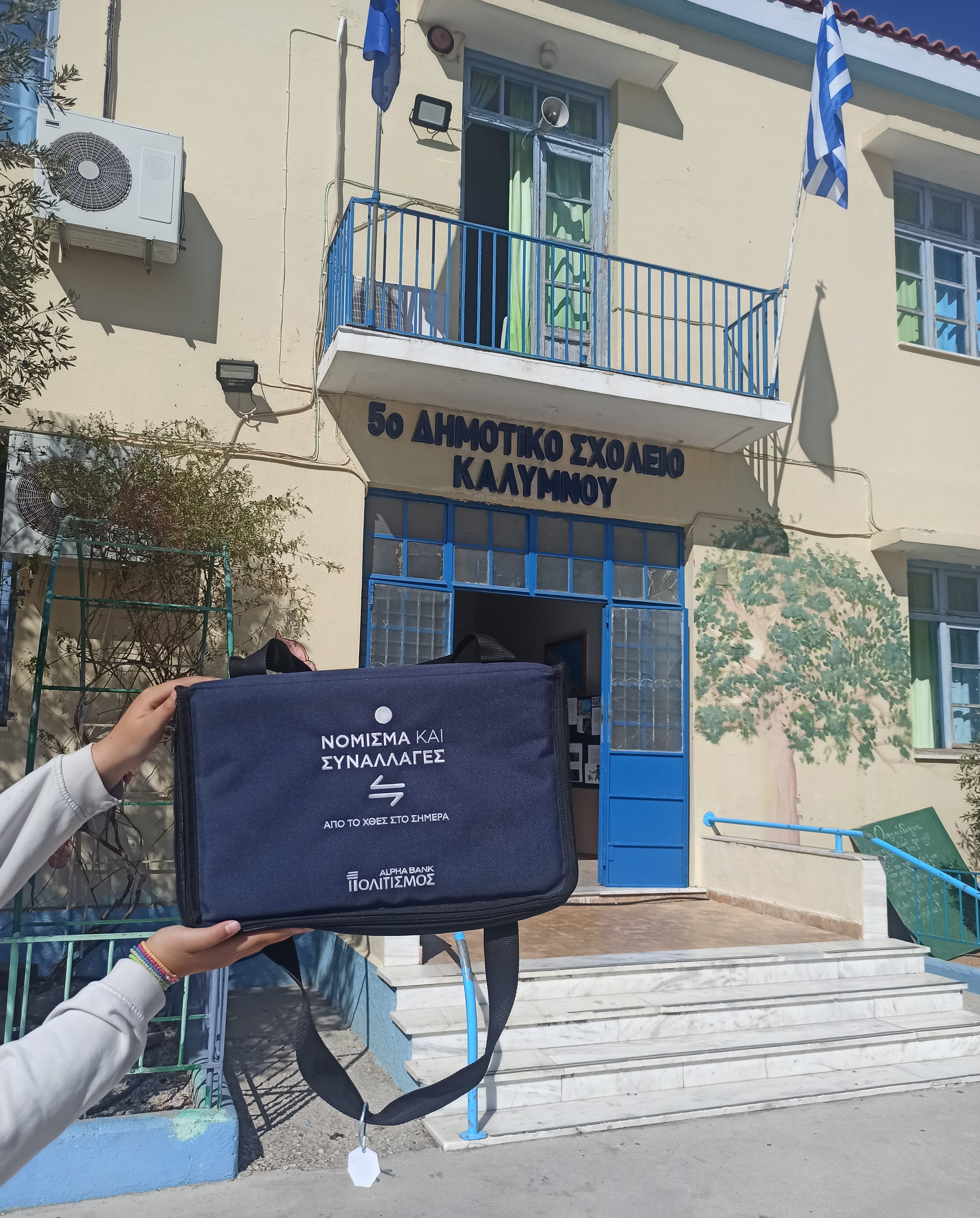 Χέρια παιδιού κρατάνε τη μουσειοβαλίτσα μπροστά από την είσοδο του 5ου Δημοτικού Σχολείου Καλύμνου. 5ο Δημοτικό Σχολείο Καλύμνου
