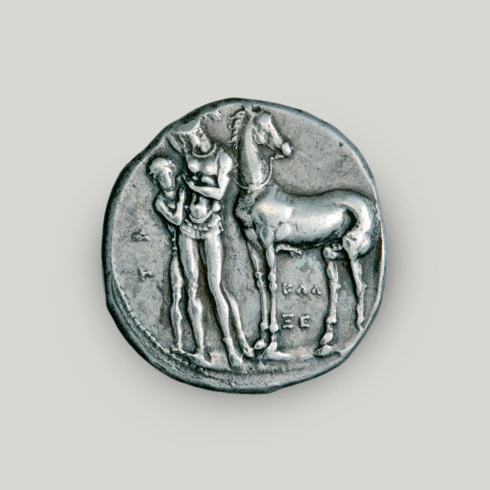Εμπρόσθια όψη αργυρού στατήρα Τάραντα με νέο δίπλα σε άλογο ενώ ο ιπποκόμος τού δένει τον χιτώνα.  Εμπρόσθια όψη αργυρού στατήρα Τάραντα