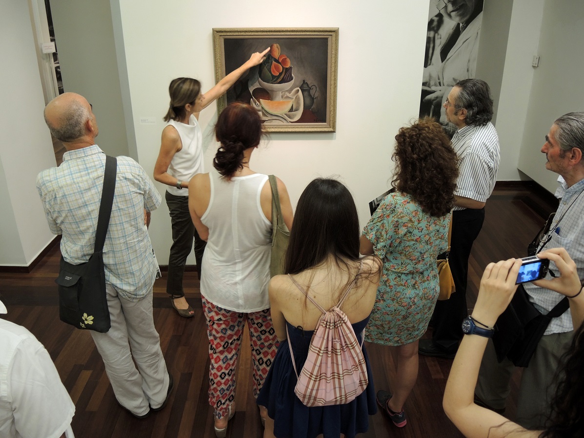 Section of the exhibition, with a picture on the wall and a tour guide describing it to the visitors around her.  Section of the exhibition “Dimitris Galanis. The Illustrated Books”