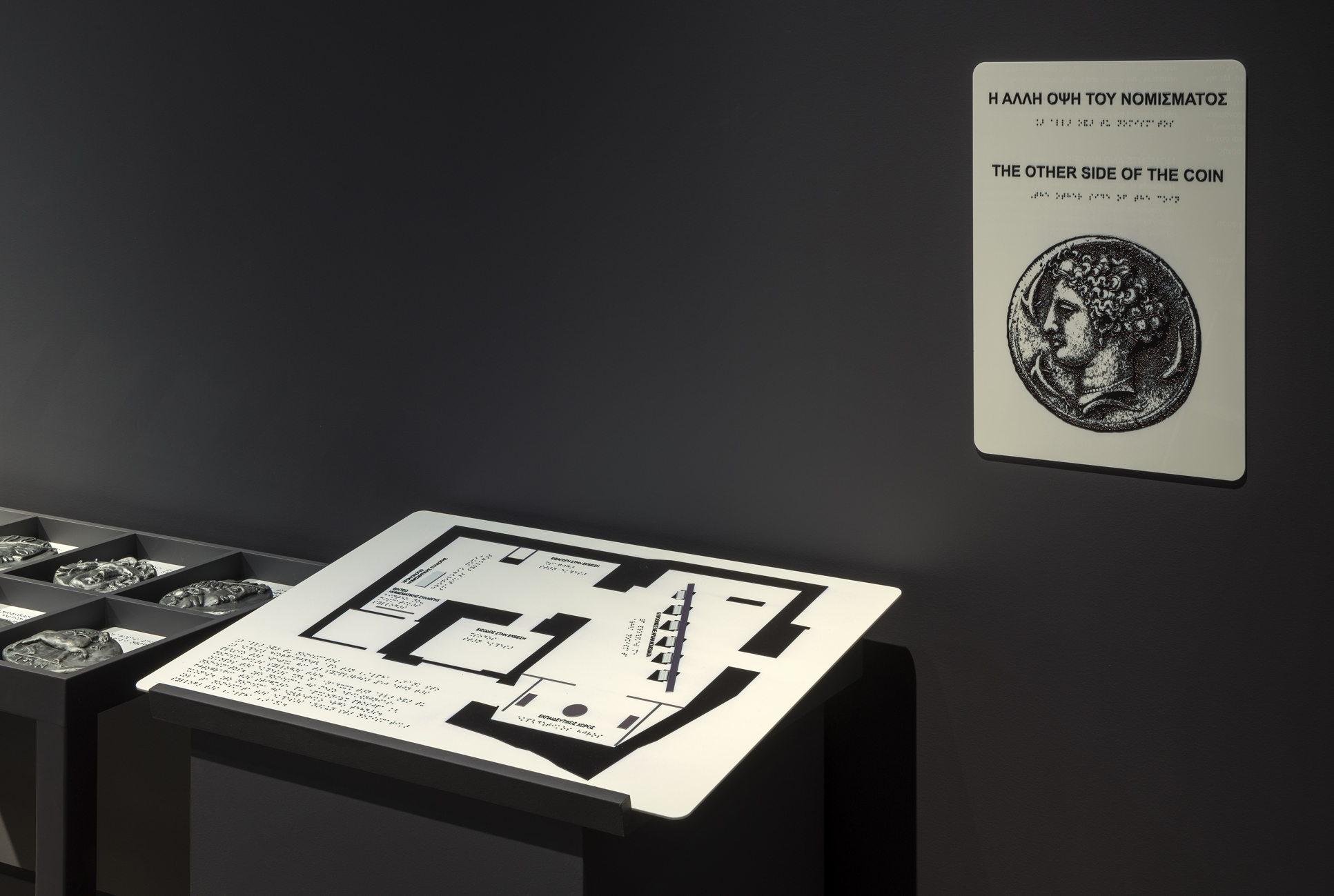 Πλακάτ σε τοίχο και επιτραπέζια επιφάνεια με πληροφορίες σε κώδικα Braille, σε Ελληνικά και Αγγλικά.  Έκθεση «Η άλλη όψη του νομίσματος»