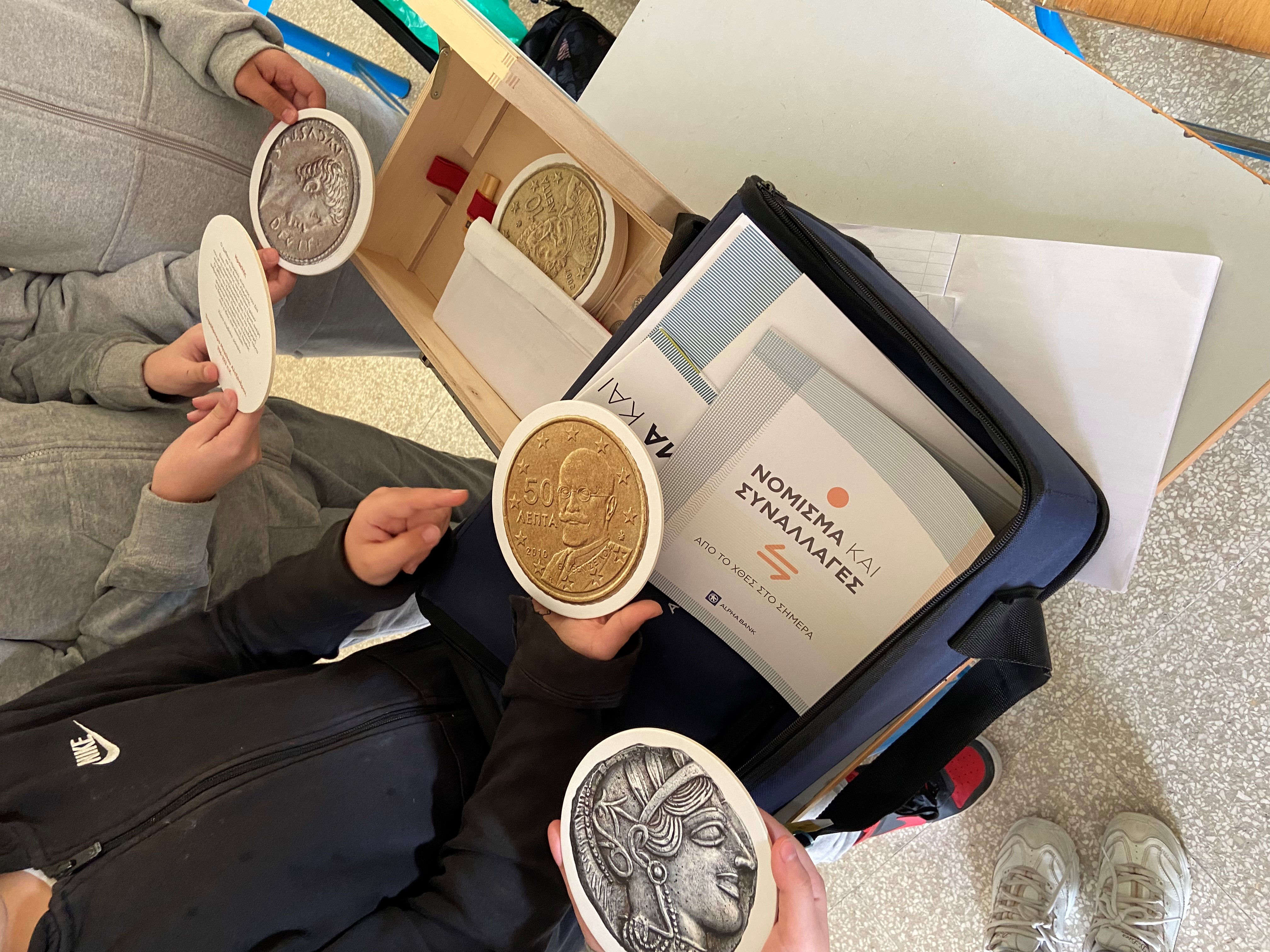 Μαθητές μπροστά από ανοιχτή μουσειοβαλίτσα με αντίγραφα νομισμάτων και φυλλάδια.  Η μουσειοβαλίτσα Νόμισμα και Συναλλαγές σε σχολείο της Κύπρου