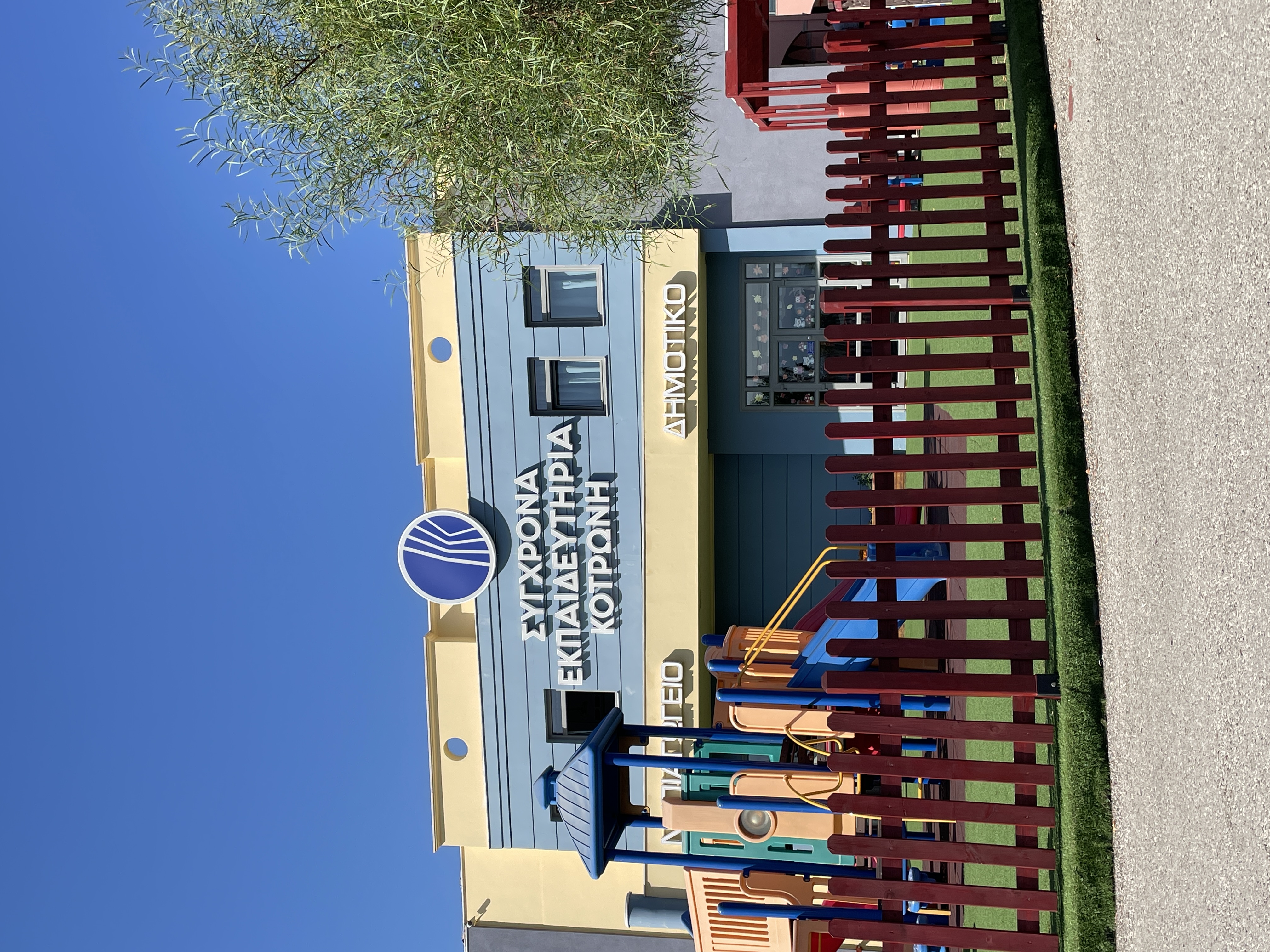 Προαύλιο με γρασίδι και τσουλήθρα και πίσω κτίριο με επιγραφή «Σύγχρονα Εκπαιδευτήρια Κοτρώνη, Νηπιαγωγείο, Δημοτικό». Πρόσοψη Εκπαιδευτηρίων Κοτρώνη στη Ναύπακτο