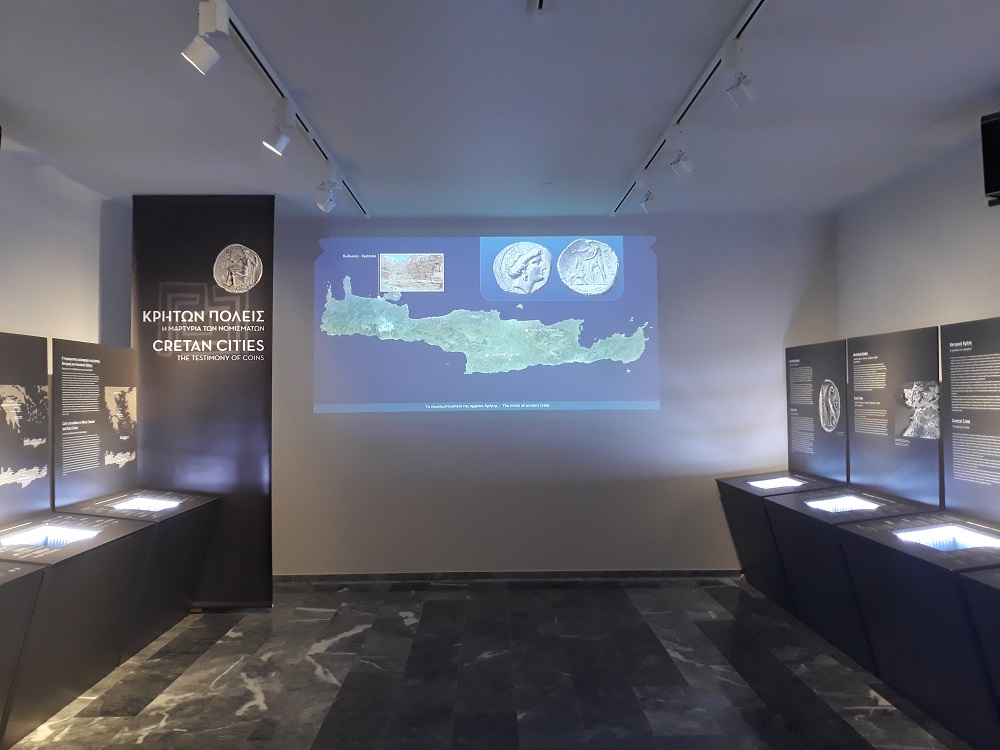 Ο χώρος της έκθεσης με εκθέματα και στον τοίχο πολυμεσική προβολή με τον χάρτη της Κρήτης.  Εκθέματα και πολυμεσική προβολή «Κρητών πόλεις. Η μαρτυρία των νομισμάτων»