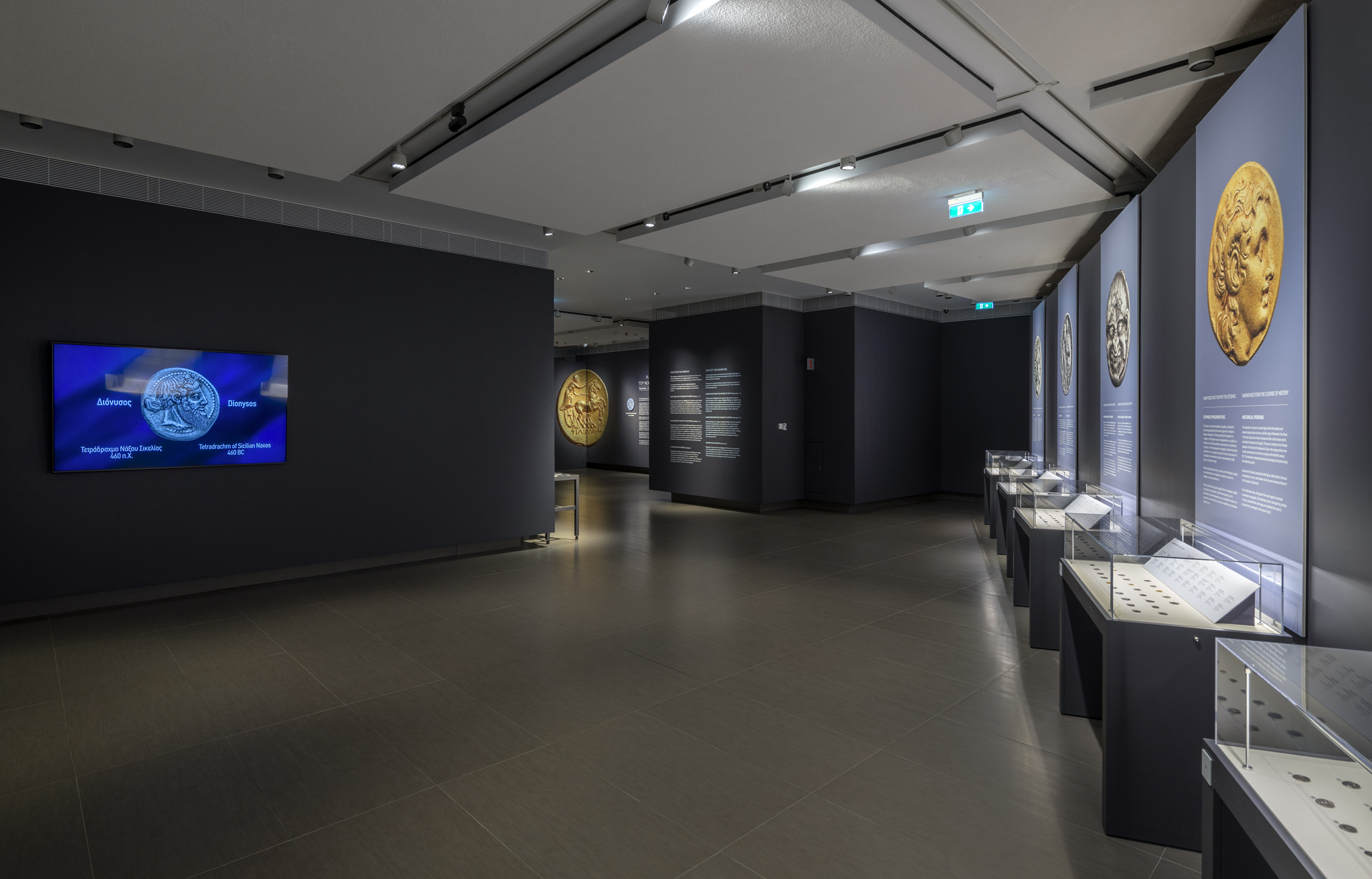 Αίθουσα με γυάλινες προθήκες νομισμάτων μπροστά από τοίχο με πληροφορίες και αριστερά οθόνη με εικόνες.  Έκθεση «Η άλλη όψη του νομίσματος»