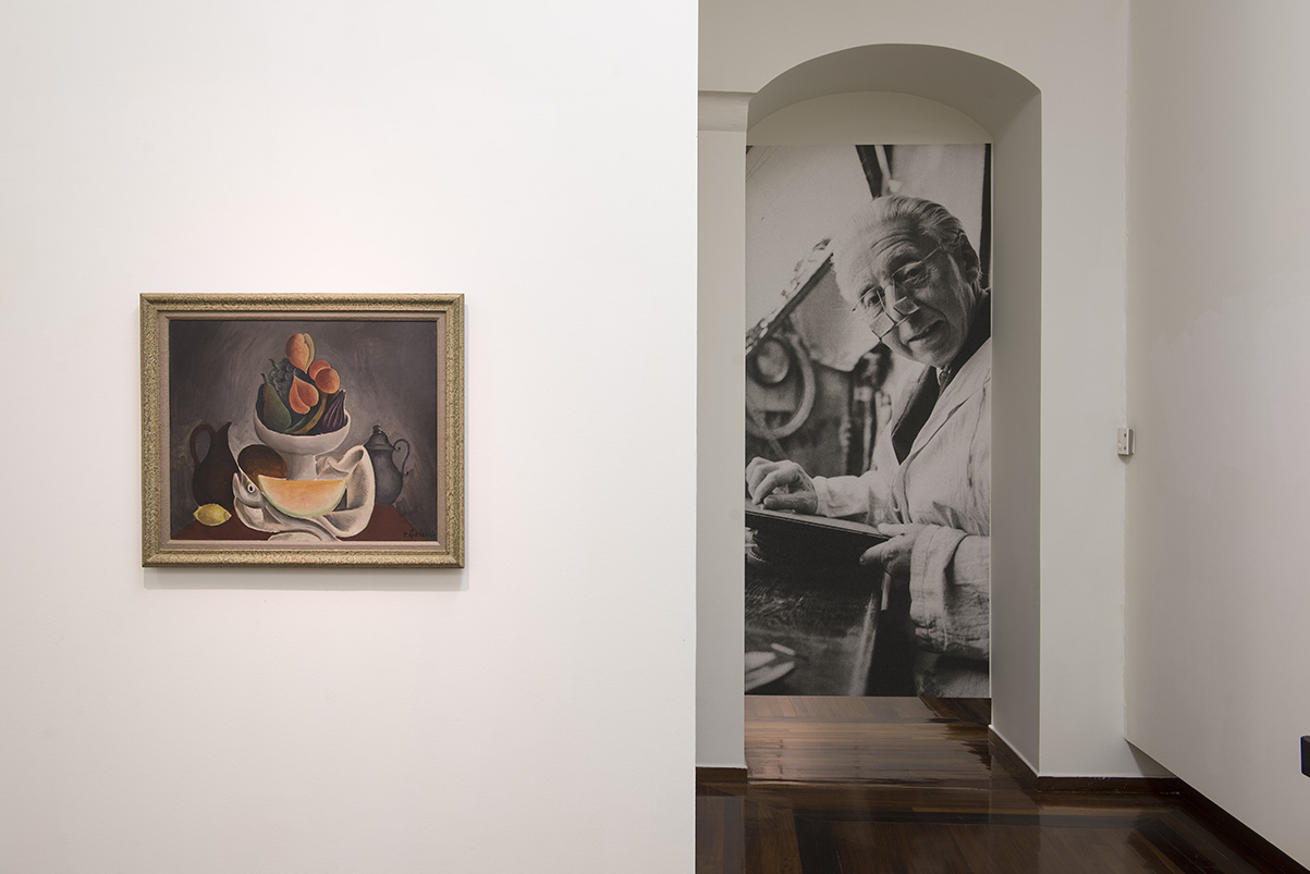 Section of the exhibition with art on the left and a large photograph of Galanis in the back.  Section of the exhibition “Dimitris Galanis. The Illustrated Books”