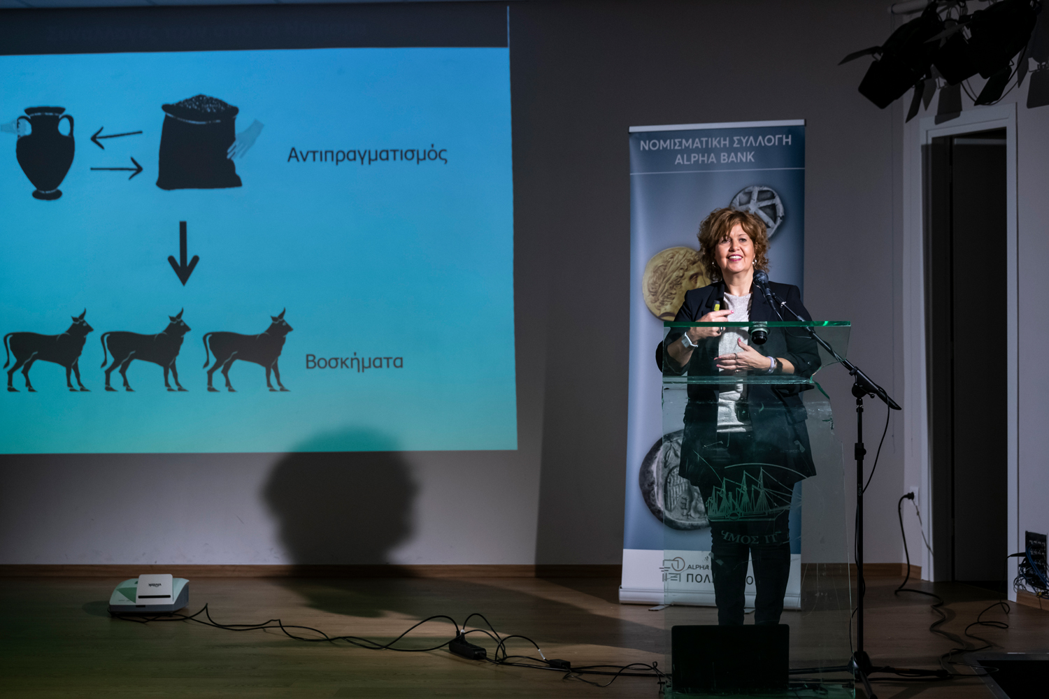 Αριστερά η Δρ Δήμητρα Τσαγκάρη στο βήμα, δεξιά παρουσίαση σε προβολέα. Παρουσίαση στο Κυριακοπούλειο Πολιτιστικό Κέντρο Ιτέας