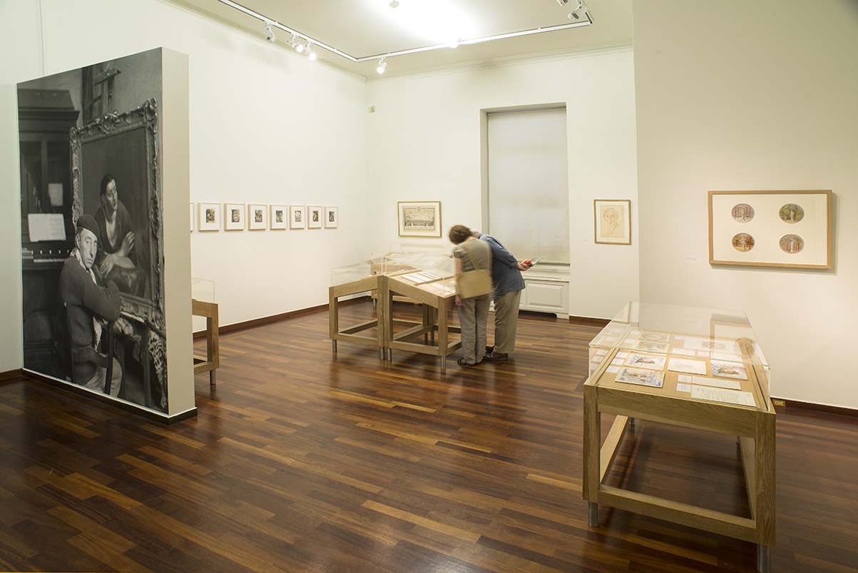 Section of the exhibition with exhibits on the walls, display cases, 2 visitors and Galanis’ photograph to the left.  Section of the exhibition “Dimitris Galanis. The Illustrated Books”