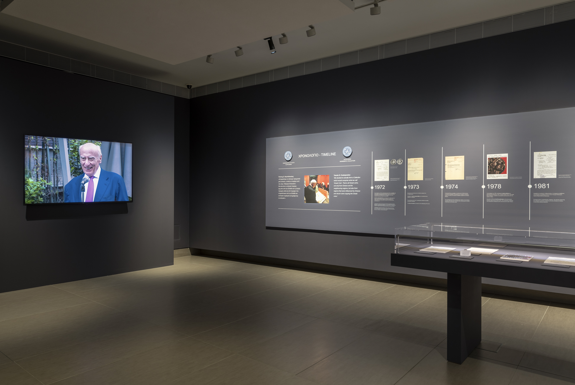 Αίθουσα της έκθεσης με γυάλινη προθήκη και τοίχο με πληροφορίες. Αριστερά οθόνη με εικόνες.  Έκθεση «Η άλλη όψη του νομίσματος»