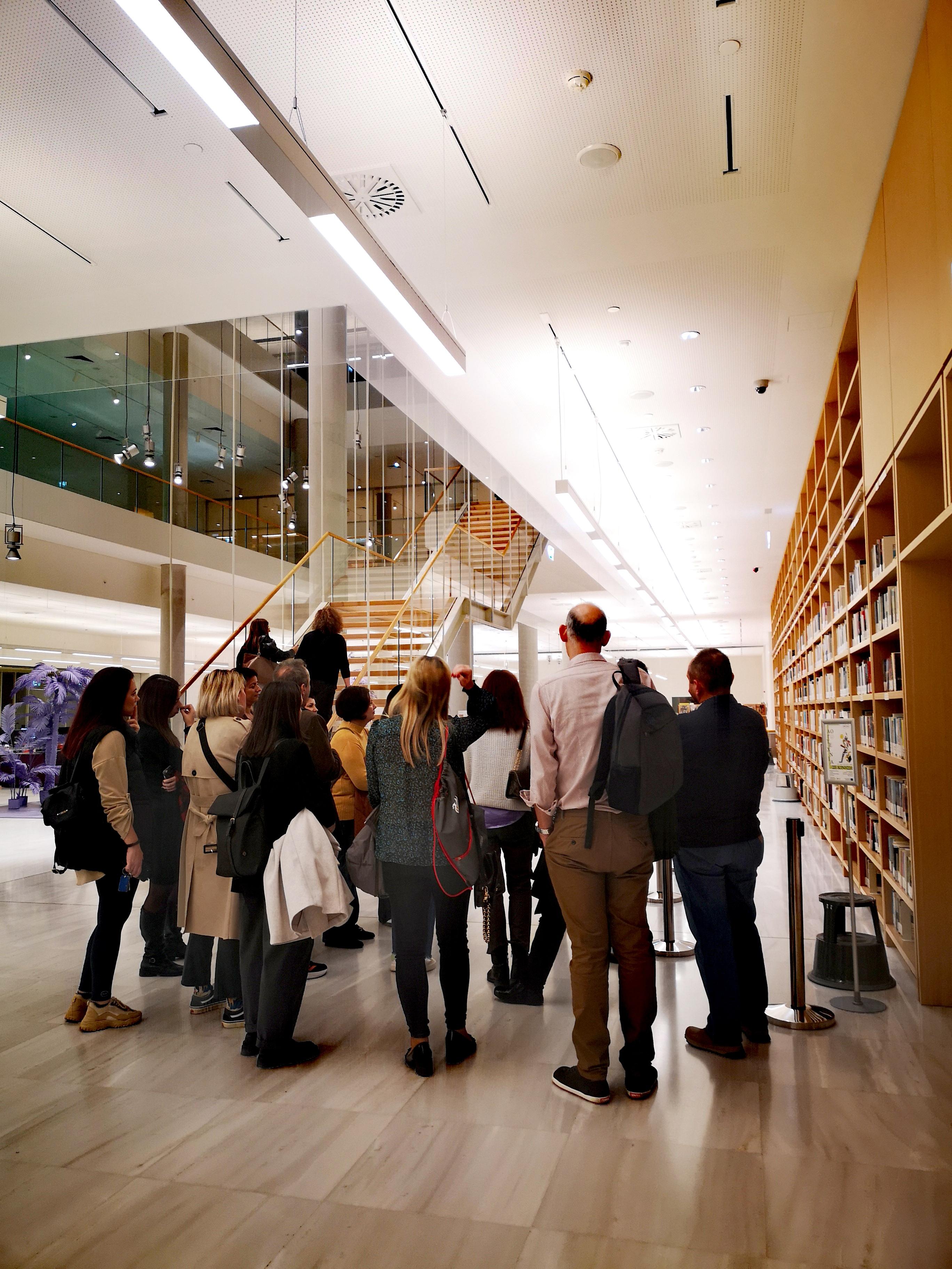Alpha Bank employees being guided through the NLG. On the right, a wall of books. In the background, a staircase. Highlight from the guided tour