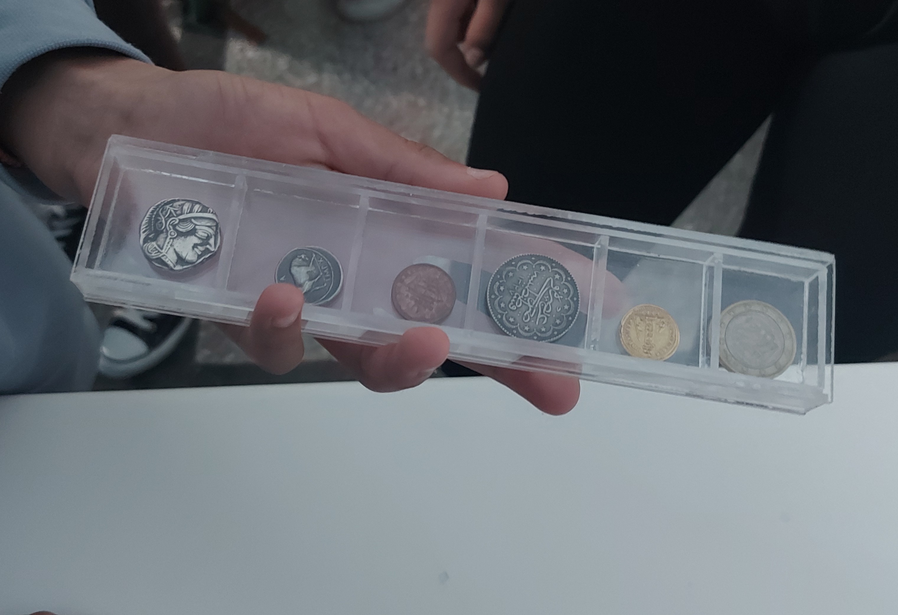 Παιδικό χέρι κρατά μακρόστενη διαφανή θήκη με 6 αντίγραφα νομισμάτων. Αντίγραφα νομισμάτων