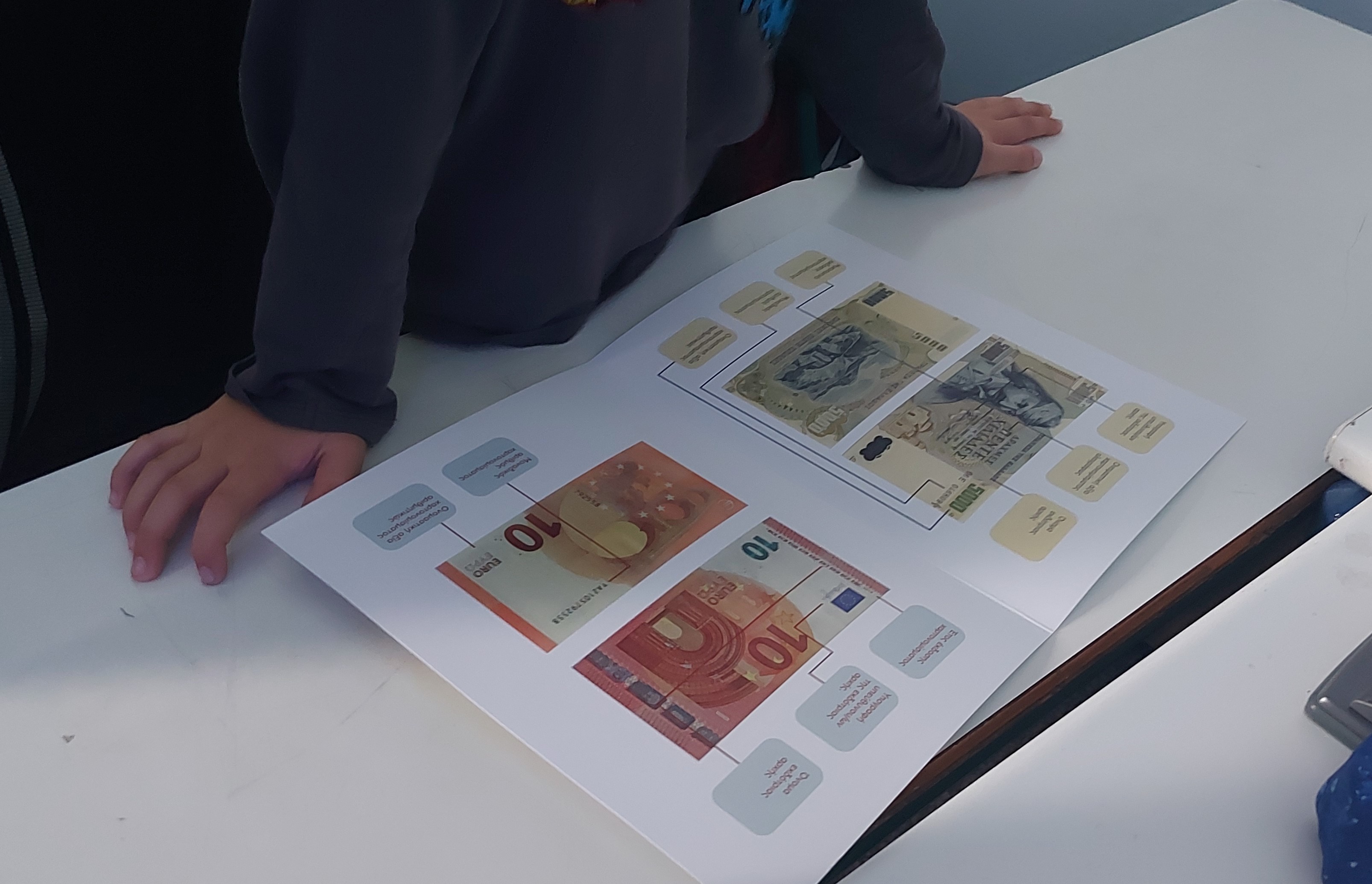 Παιδί σε θρανίο κοιτά έντυπο που εξηγεί τη μορφολογία ενός χαρτονομίσματος 5.000 δραχμών και ενός 10 ευρώ. Εκπαιδευτικό υλικό