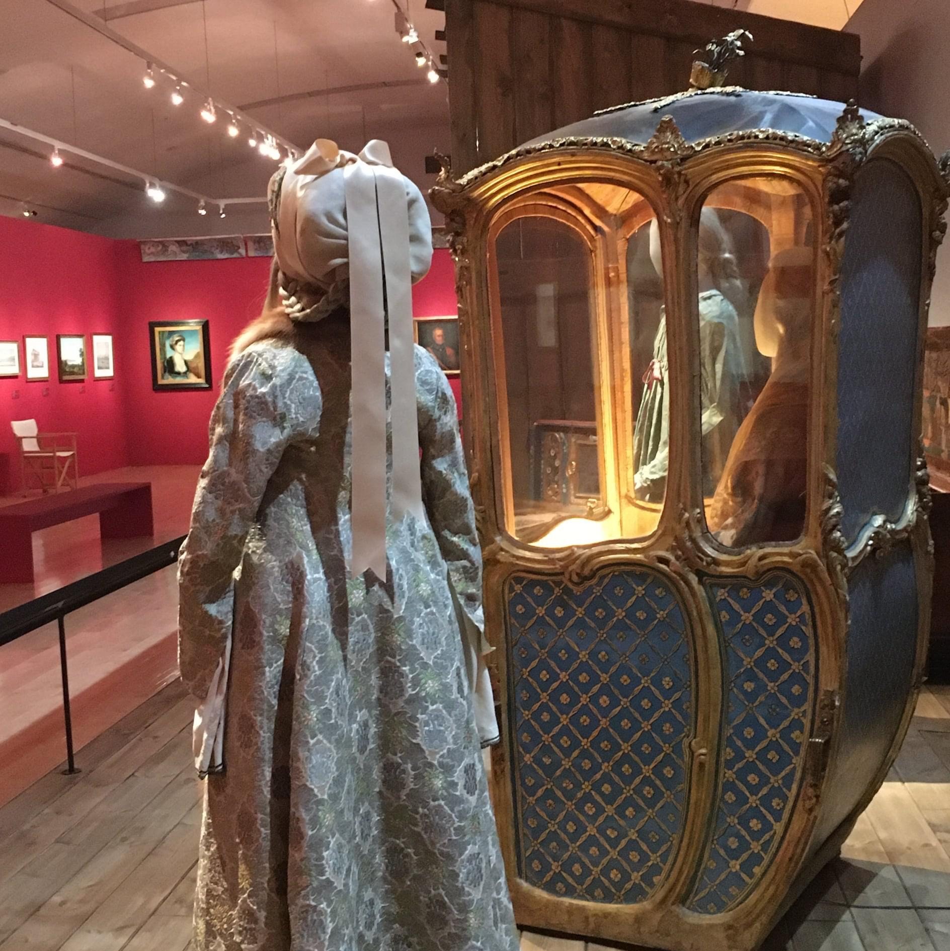 3D figures of women and a small carriage. Paintings hanging at the back corner of the room. Exhibition “1821 Before and After”