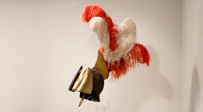 Μπρούντζινη περικεφαλαία με φτερά, μπροστά από λευκό τοίχο πάνω σε διάφανη βάση. 