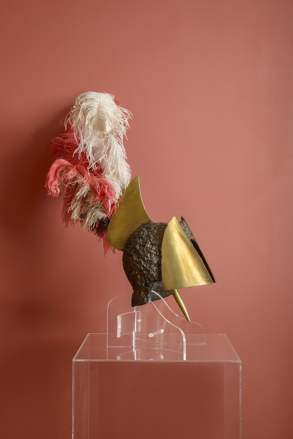 Μπρούντζινη περικεφαλαία με φτερά, μπροστά από κόκκινο τοίχο πάνω σε διάφανη βάση. 