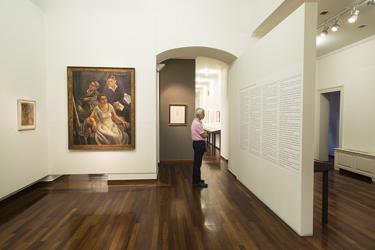 Section of the exhibition, with a painting on the facing wall, text on the wall to the right and a visitor.  Section of the exhibition “Dimitris Galanis. The Illustrated Books”